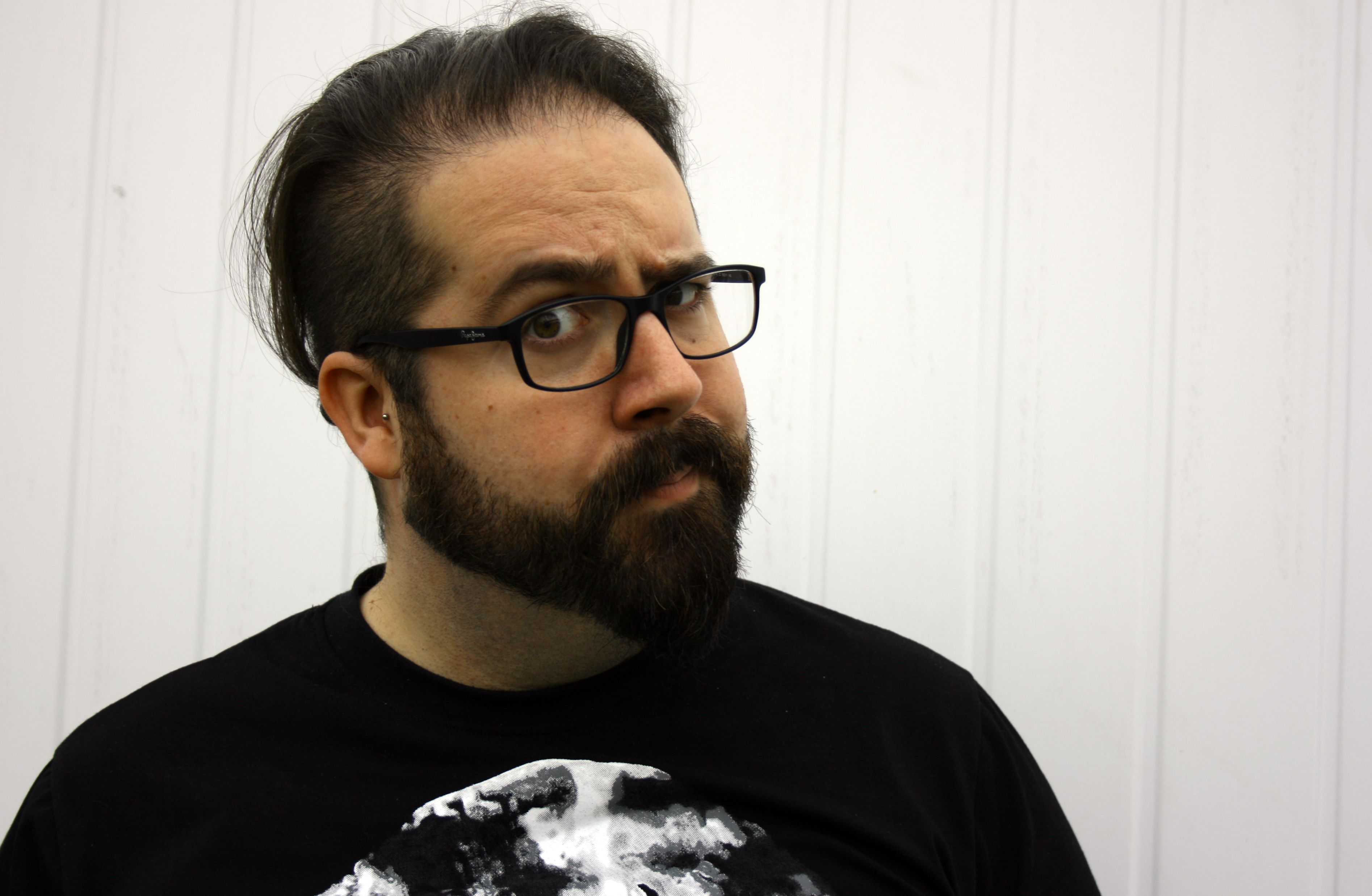 The head and shoulders of a white man with short black hair, and a short, bushy black beard and mustache. He is wearing square black glasses and is raising one eyebrow quizzically at the camera.
